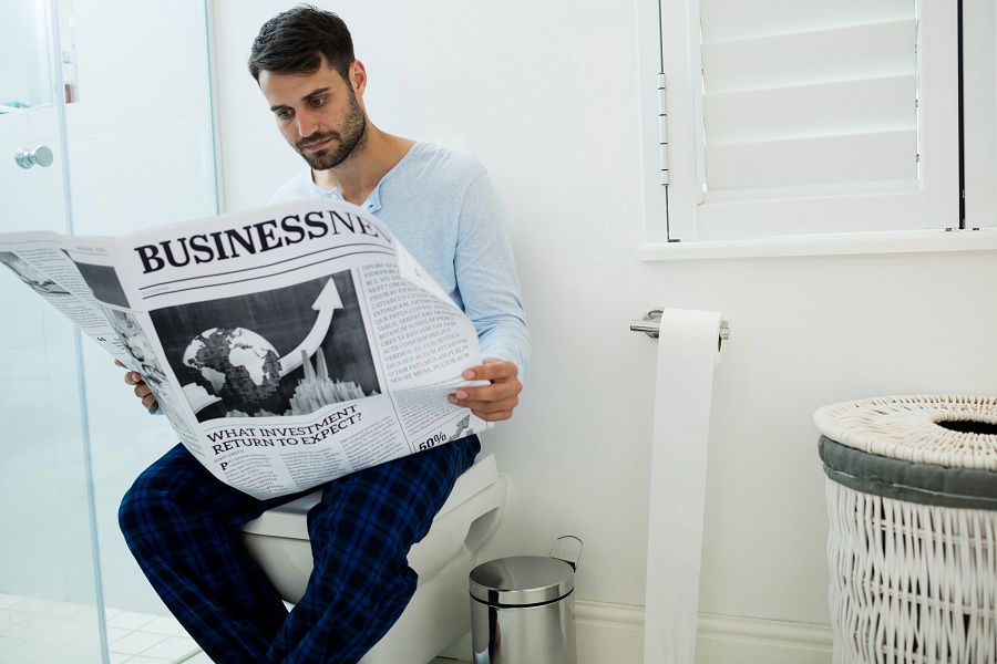 man sitting on toilet while reading a newspaper in the morning